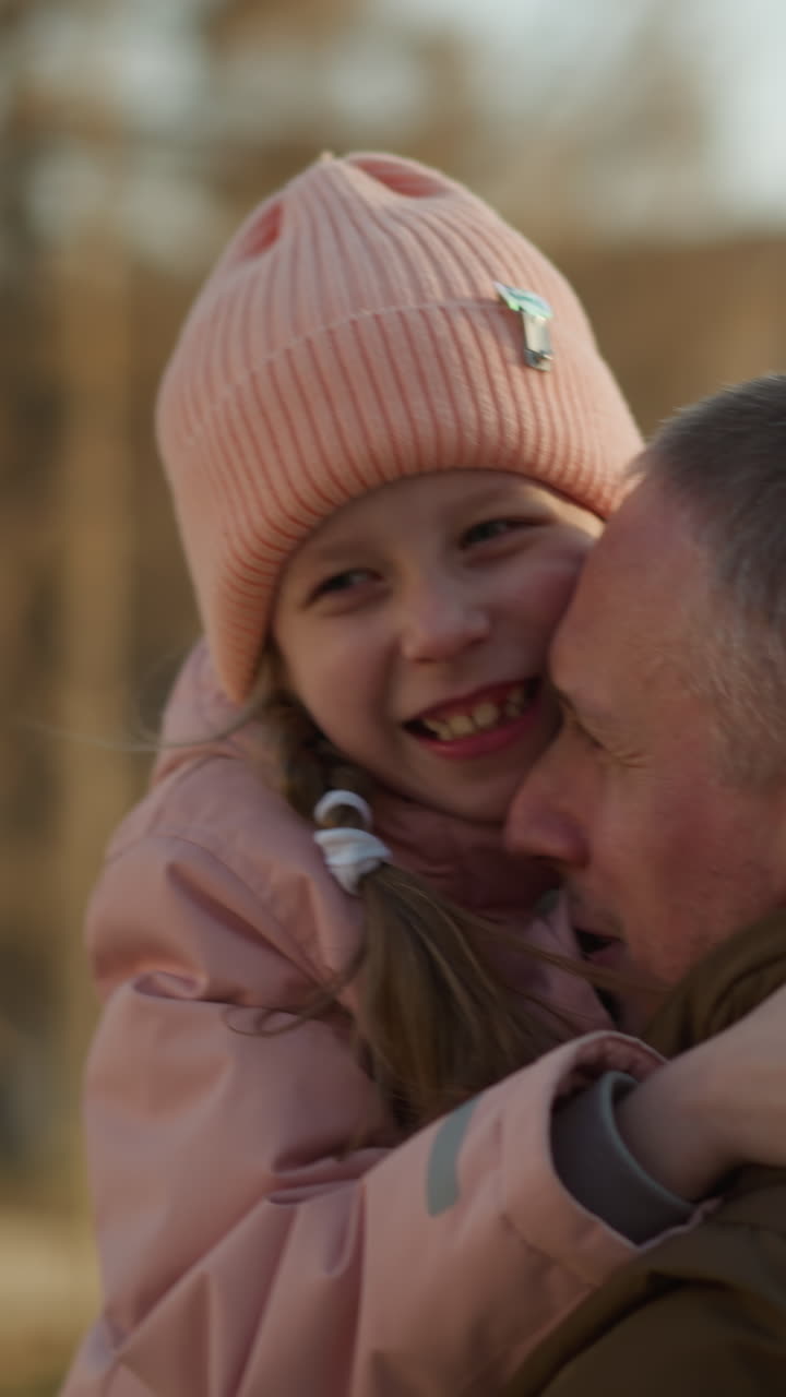un hombre con una chaqueta marrón lleva a una niña con una gorra rosada y una chaqueta mientras ella lo abraza fuertemente. la escena captura un momento tierno y amoroso de unión padre-hija al aire libre en un día soleado
