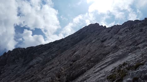 back-view-of-a-climber-with-orange-helmet-ascending-a-german-via-ferrata-to-the-mountain-Alpspitze,-securing-himself-on-the-steel-cables,-camera-pan-right-to-left-and-back