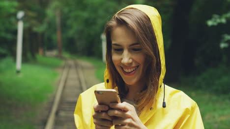 Close-Up-Of-Cute-Woman-In-A-Yellow-Raincoat-Standing-In-The-Middle-Of-The-Woods,-Laughing-And-Texting-On-Her-Smartphone