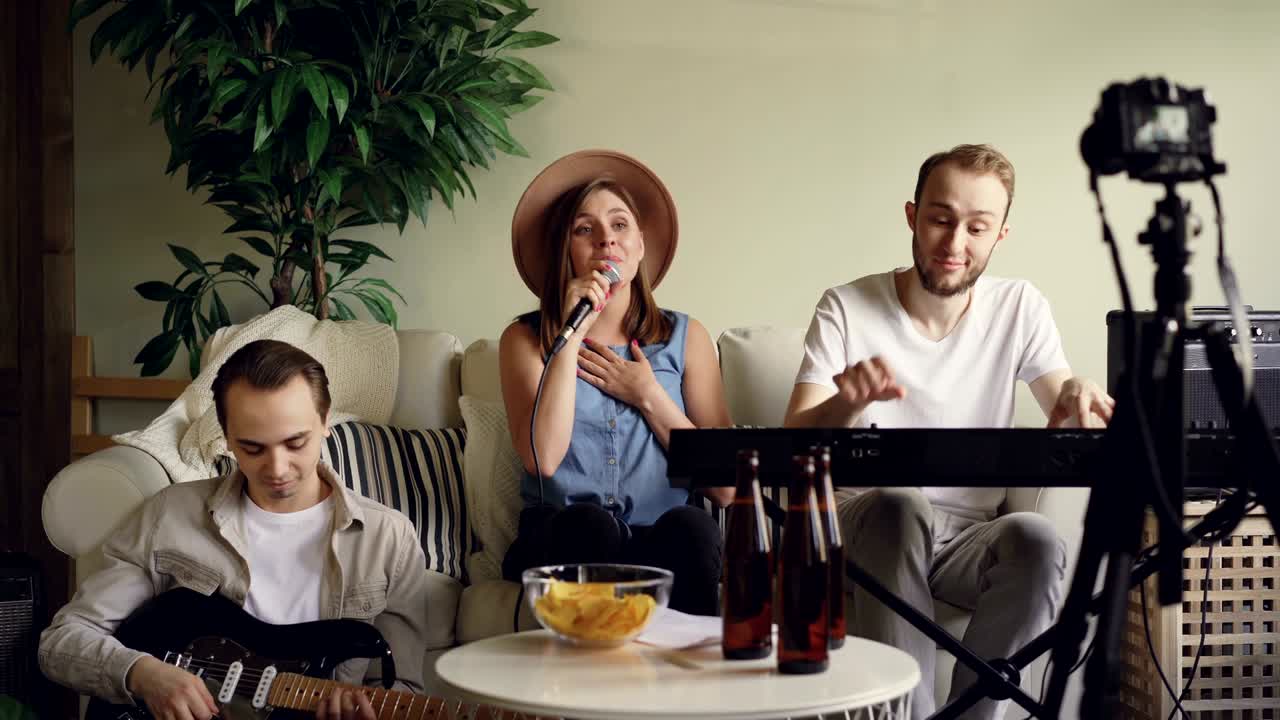 el grupo musical está practicando en casa y grabando su práctica en cámara para su blog en línea en internet. el vocalista está cantando, el tecladista y el guitarrista están tocando.