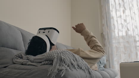 man lies on couch with blanket using vr headset and raising hand to interact