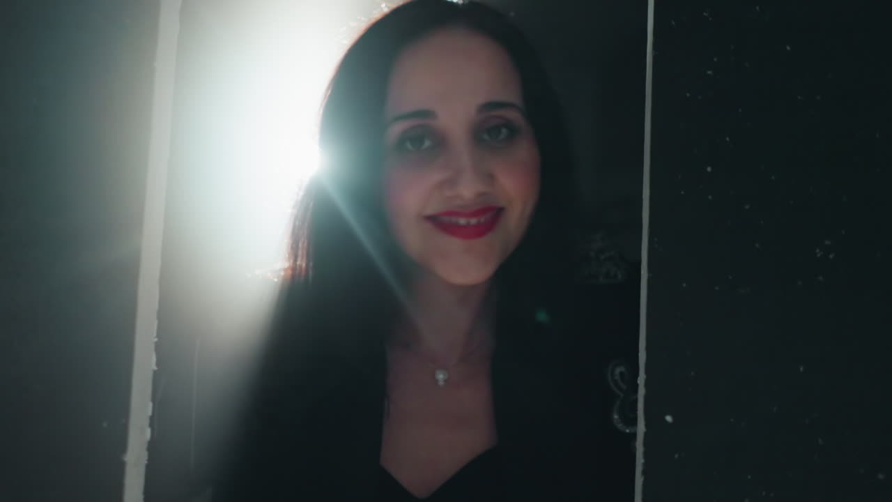 Intimate close-up of a woman in black with red lipstick, smiling gently as the camera zooms in and light flares dramatically
