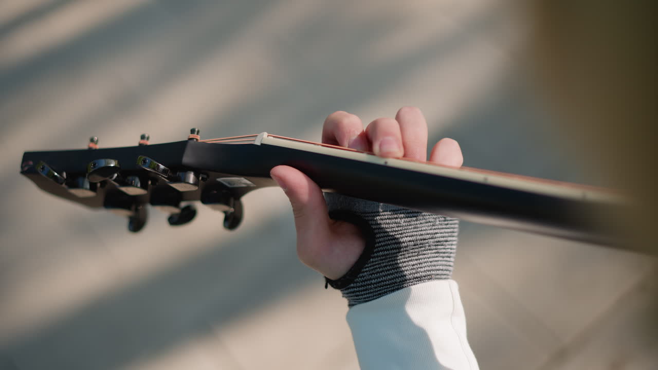 left hand on guitar neck, closeup of lefthanded guitarist wearing grey knitted glove, sunlight and shadow patterns across fretboard, slowmotion fretting and tuning peg detail in quiet urban