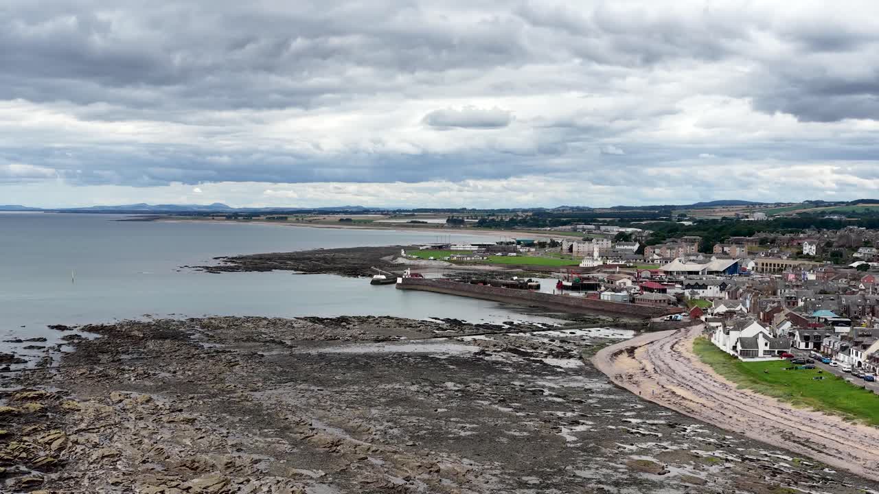 Luftaufnahmen mit Kameraschwenk über felsige Küstenlinie, Hafen und Stadt unter bewölktem Tageslicht in Dundee, Schottland
