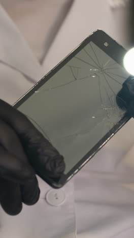 una vista de cerca de un técnico en una bata de laboratorio blanca y guantes negros examinando una pantalla de teléfono inteligente agrietada