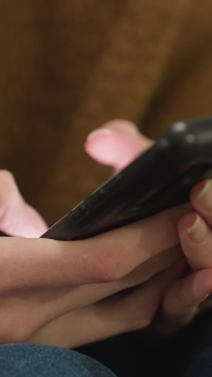 Close-up of hands wearing a brown coat and blue jeans, holding and pressing a smartphone with both hands. the interaction with technology, emphasizing the focus on the device in an urban setting