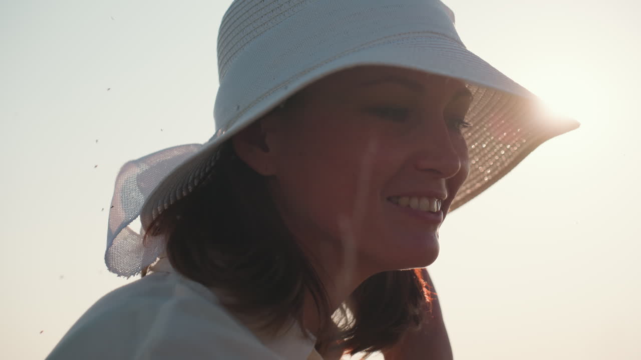 lady in wide hat smiling gently while admiring something in warm sunlight, insects fly around adding movement, soft morning glow outlines hat creating serene and intimate summer