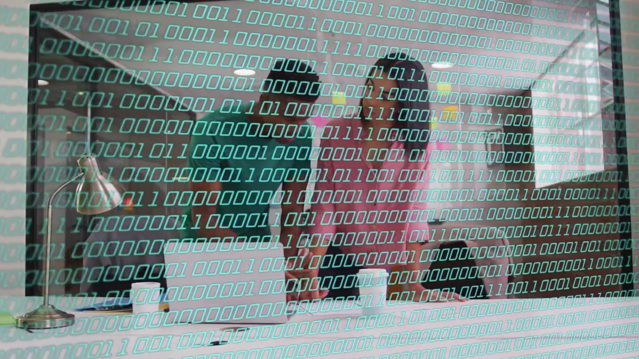 Team members leaning over laptop, reviewing tech data, teal binary overlay revealing sticky notes