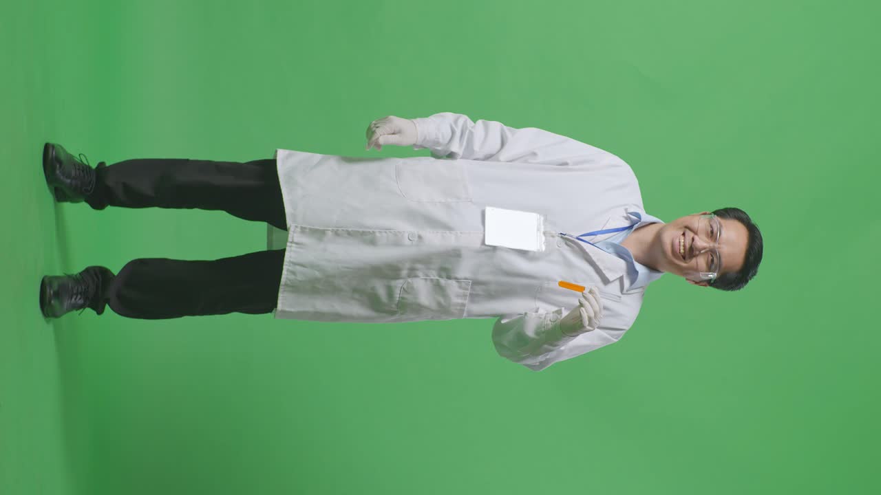 Full Body Of Asian Man Scientist Smiling And Showing Orange Liquid In The Test Tube To Camera While Standing On The Green Screen Background In The Laboratory
