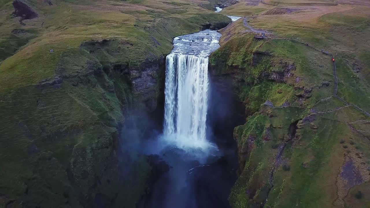 Breathtaking aerial view of Skógafoss waterfall plunging down a cliff face, surrounded by lush green landscape in Iceland, creating a mesmerizing natural wonder, drone pull out establshing shot