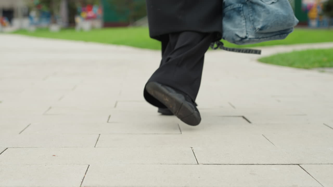 giovane studente che cammina nel campus, primi piani dal basso con angolo ridotto di scarpe da ginnastica nere e pantaloni ampi, zaino di jeans che oscilla, passo svelto sul vialetto pavimentato, parco urbano leggermente sfocato, movimento spontaneo