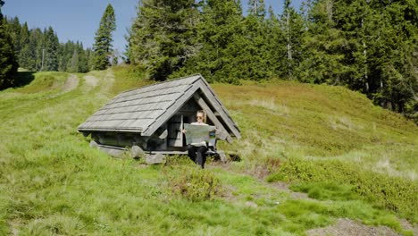 young woman reads unfolded map, sitting in front of small old wooden mountain hut, placed in the middle of mountain clearing