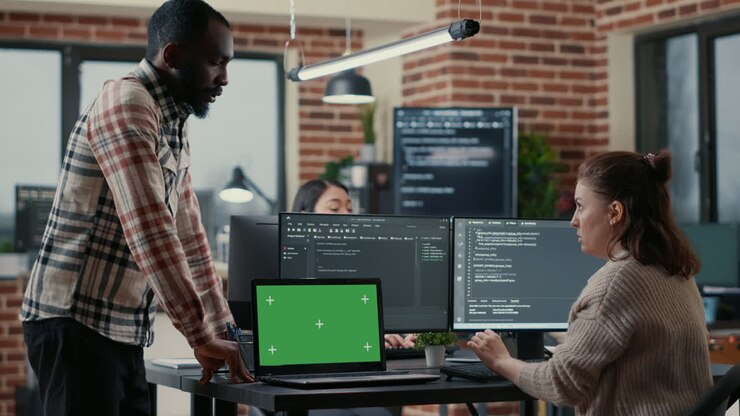 Programer working at desk with multiple computer screens and laptop with green screen chroma key mockup