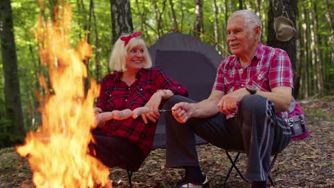 una vieja pareja feliz de abuelos descansando en el campamento en el bosque cocinando salchichas fritas sobre la fogata