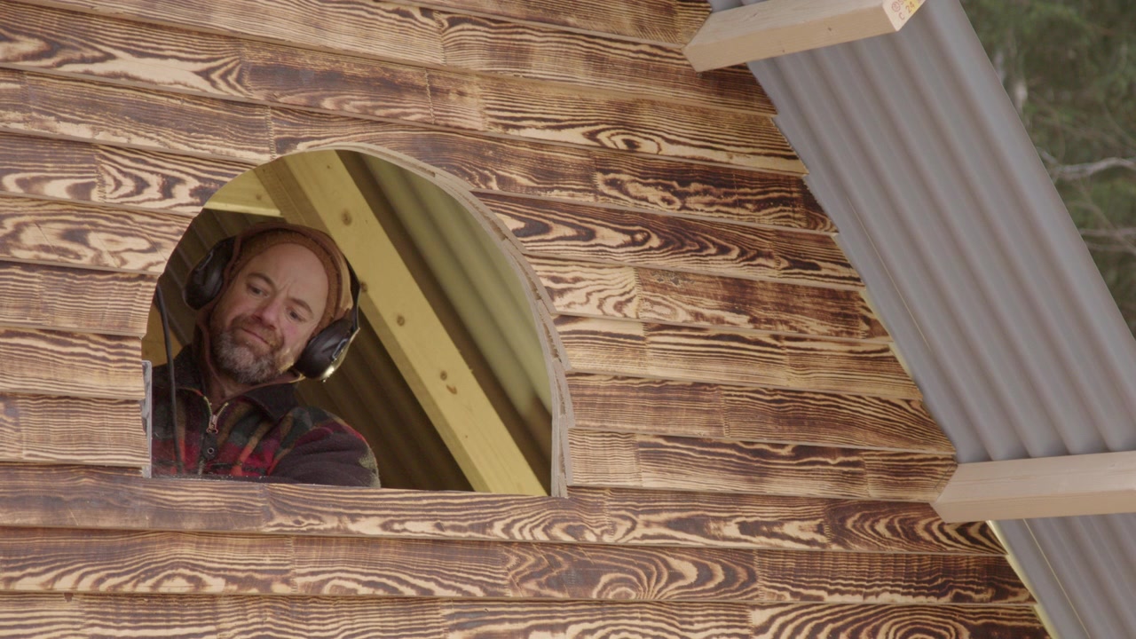 Tuttofare con cuffie antirumore di sicurezza che taglia una nuova finestra in una piccola baita di legno, teleobiettivo