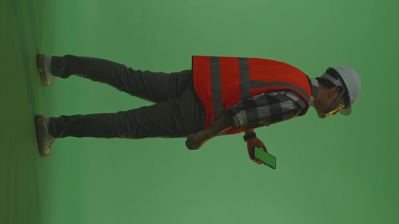 Full Body Back View Of Asian Male Engineer With Safety Helmet Working On A Green Screen Smartphone And Looking Around While Standing In The Green Screen Background Studio, During Sunset Or Sunrise Time