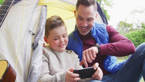 Feliz-Padre-Caucásico-Con-Su-Hijo-Sentado-En-Una-Tienda-Y-Usando-Un-Teléfono-Inteligente-En-El-Jardín