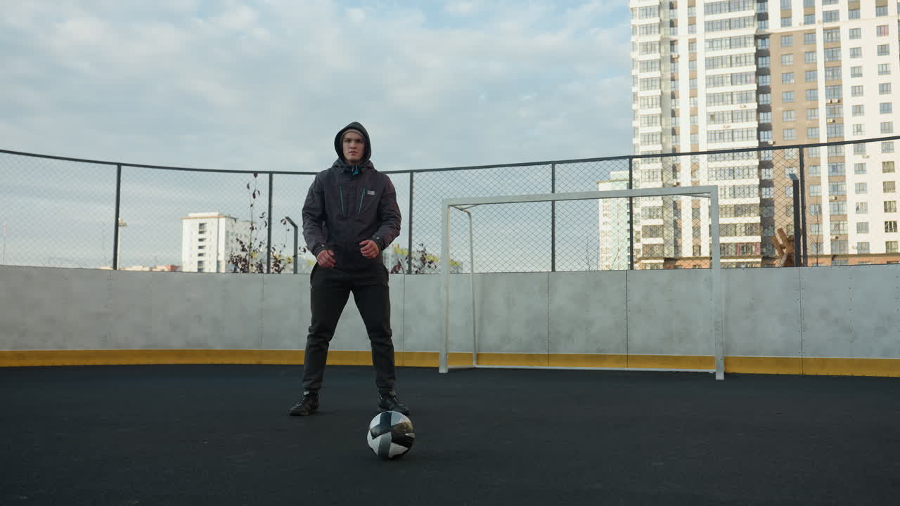 entrenador realizando sentadillas con las manos juntas en preparación para el ejercicio en un campo deportivo al aire libre, con la pelota de fútbol en primer plano, rodeado de edificios residenciales urbanos de gran altura y postes de gol