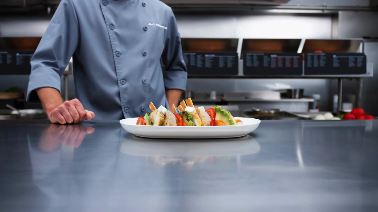 La telecamera si avvicina a un piatto da portata bianco riempito con cibo ben disposto, verdure a foglia e peperone rosso, mentre uno chef in uniforme rimane dietro il bancone tra gli arredi da cucina in acciaio inossidabile