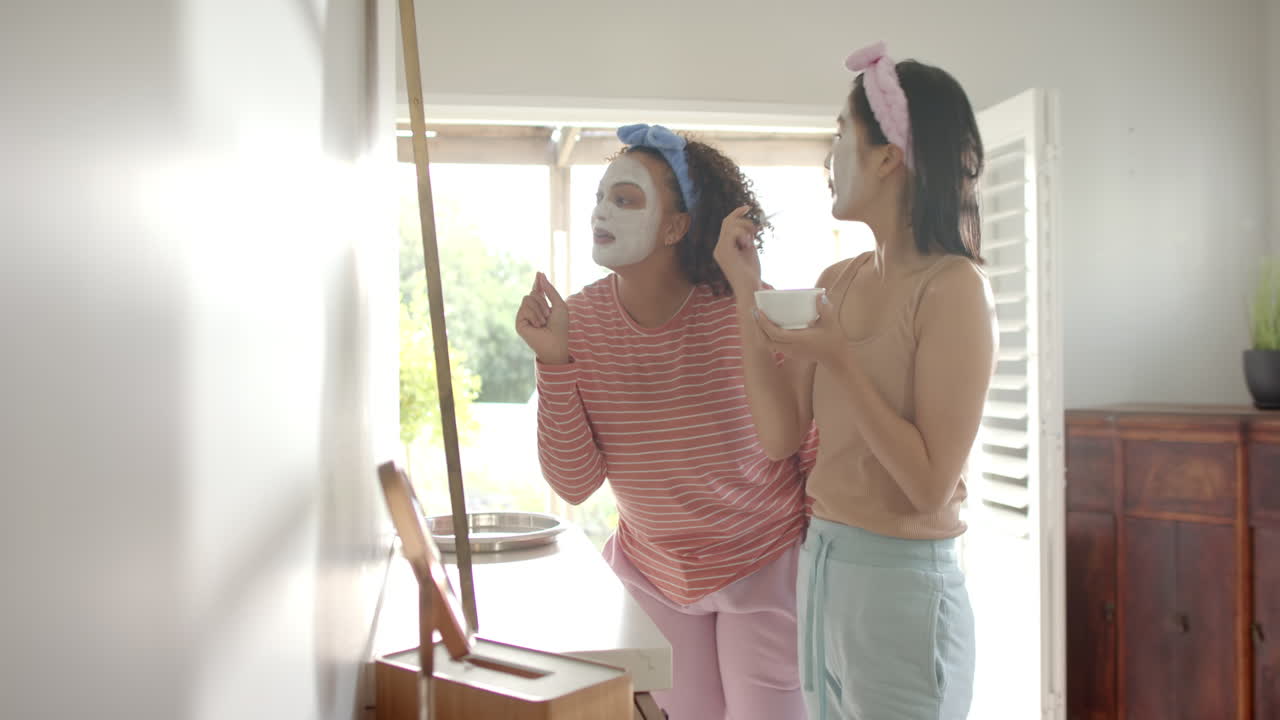 Applying face masks, two diverse women friends enjoying skincare routine at home together