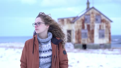 young girl looks around in a snowy environment, looking lost. focus switches from her face to a rusty house in the background. close up stable shot