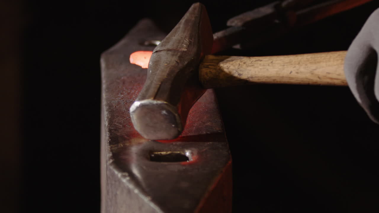 Ironsmith's gloved hand picks up hammer to forge red-hot iron on anvil, close-up
