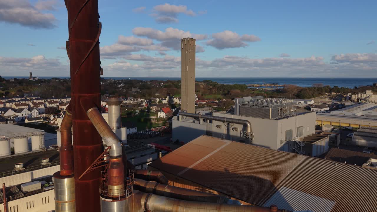 Imagens de drone de St Sampson, Guernsey, mostrando um complexo de central elétrica e chaminés, passando perto da chaminé sob o sol dourado com um céu e mar interessantes ao fundo