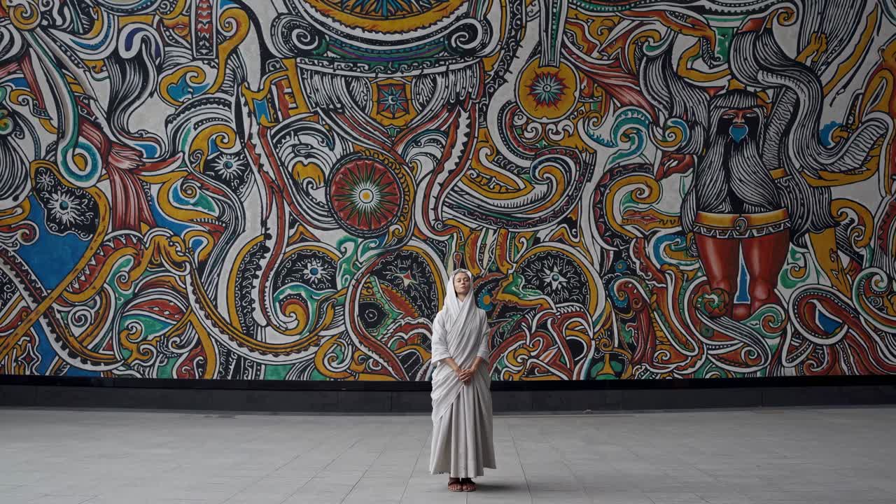 Woman wearing a religious habit meditating in front of a large mural representing norse mythology, creating a contrast between spirituality and ancient legends