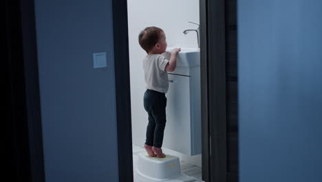caucasian toddler boy stands on the prop near the sink in the bathroom. happy baby opens and closes the faucet.