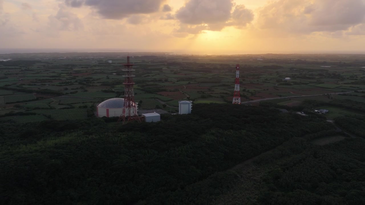 Horisontale luftoptagelser, der bevæger sig til venstre og derefter panorerer til højre over kommunikationstårne og det omkringliggende landskab under solnedgang på Irabu-øen, Miyako, Okinawa, Japan.