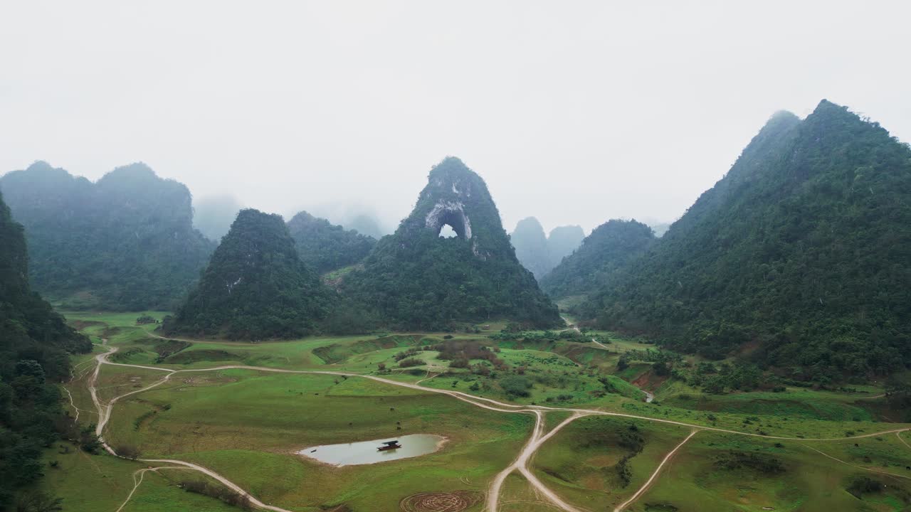Núi đá vôi karst với vòm đá tự nhiên bị che khuất một phần bởi sương mù dày đặc và mưa nhẹ, tạo ra bầu không khí u ám trong tầm nhìn thấp từ góc nhìn drone trên không tại Núi Mắt Thần Việt Nam.