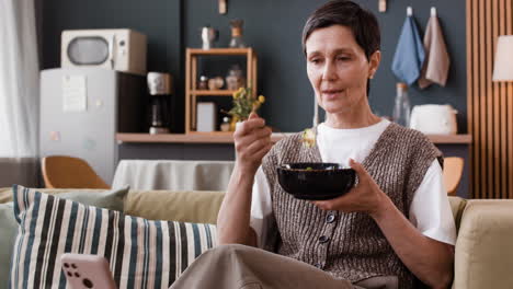 woman eating a meal on a couch at home while looking at a smartphone