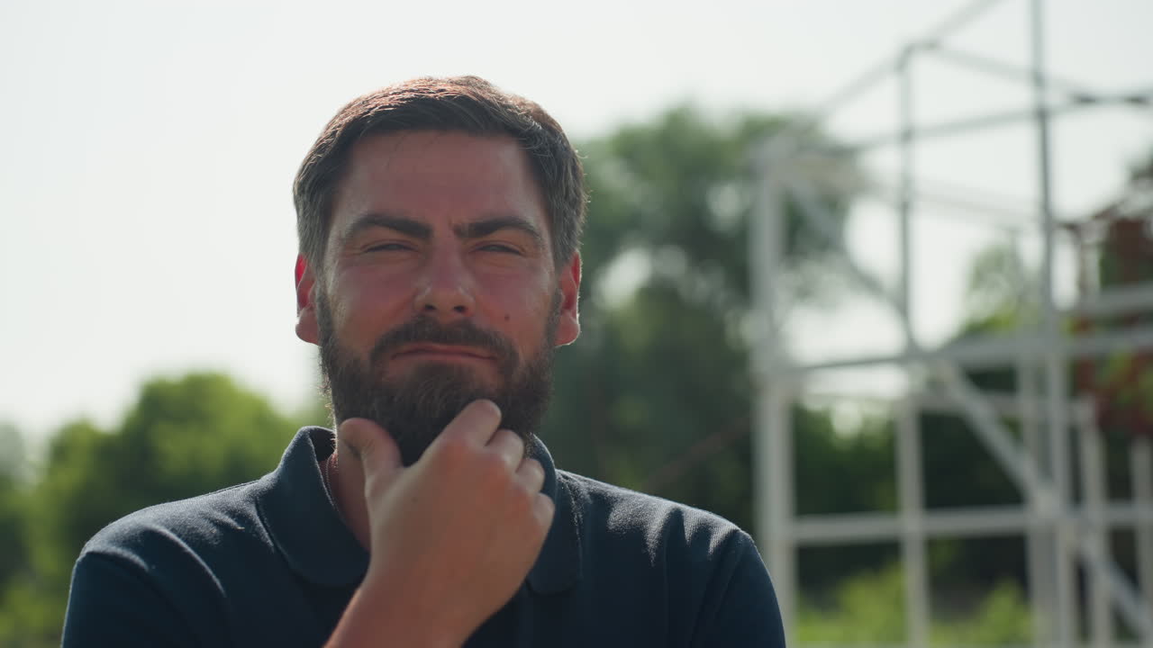 young man touching beard thoughtfully while gazing into distance near metal frame structure calm expression suggesting reflection and decision making on rural project