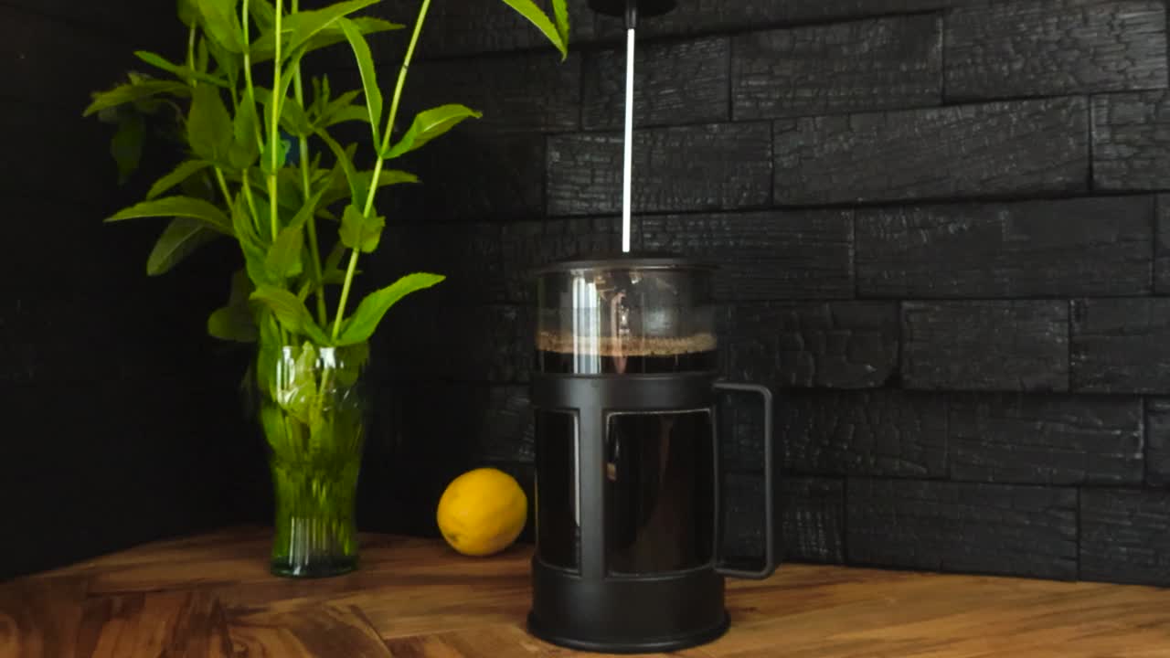 Close up kitchen scene, French coffee press on a warm olive wood table with mint stems in glass vase and lemon on countertop. Backdrop of yakisugi charred black wood panels. Cozy coffee making at home