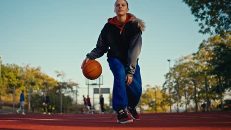 below is a portrait of a blonde girl in a sports uniform and blue pants who is hitting an orange basketball ball off the red floor on a street court in the summer