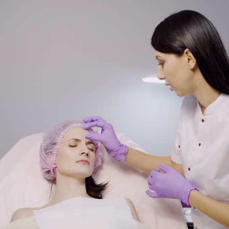 A beautiful cosmetologist advises a client who lays on a chair and examines her facial wrinkles in the aesthetic beauty salon on a gray background.