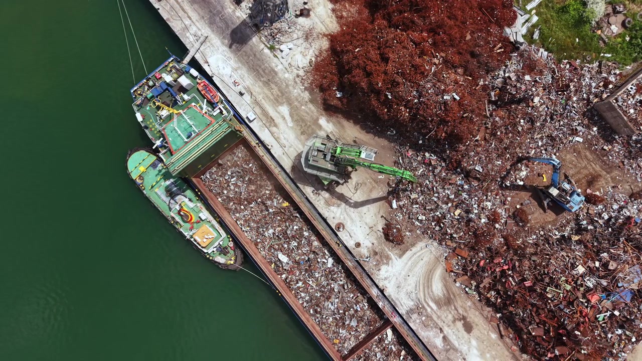 Nave scarica rottami metallici in un cantiere di riciclaggio situato vicino a un fiume di giorno, mentre gru e veicoli lavorano nelle vicinanze per spostare materiali e macchinari.