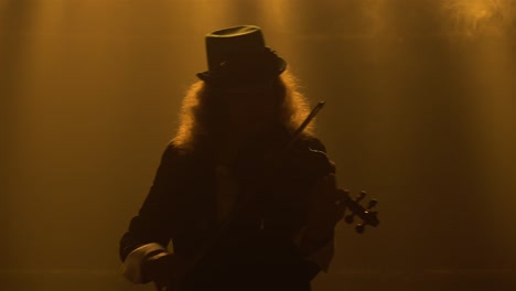 silhouette of a violinist musician with lush hair wearing a hat plays a classical wooden violin. a man performs a recital in a dark smoky studio with yellow lights. close up