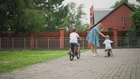 a woman happily reaches out to her younger child riding a bicycle, offering support and encouragement, the older sibling rides from behind, with a building behind them