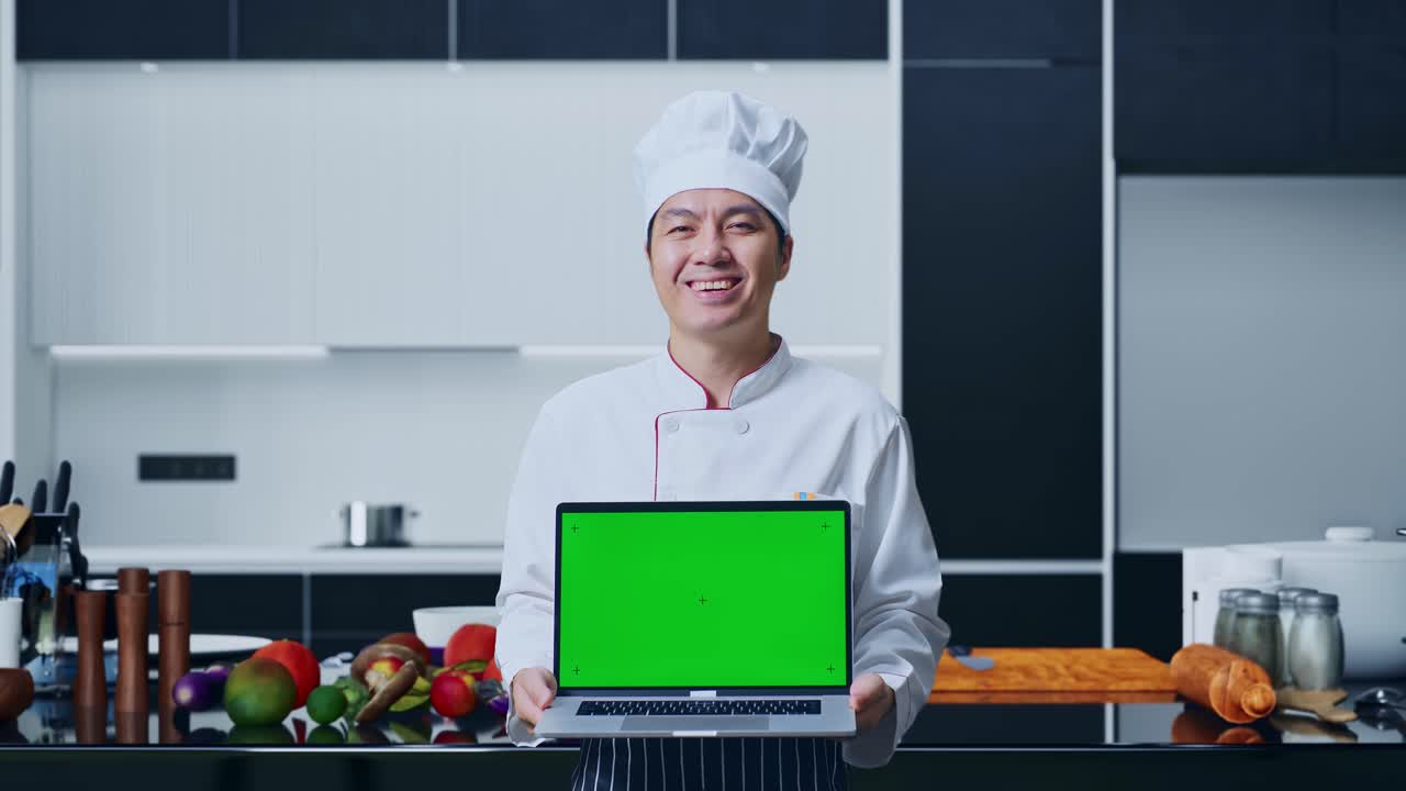 아시아인 셰프가 미소 짓고 집 주방에 서 있는 동안 카메라에 녹색 화면 노트북을 보여줍니다.