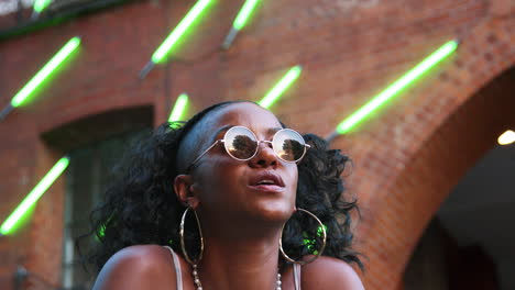jovem negra à moda usando óculos de sol redondos sentada na rua, ângulo baixo, close-up, luzes de faixa de néon desfocadas no fundo