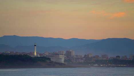 Filmisch-Atemberaubend-Noch-Nahaufnahme-Dramatische-Leuchtend-Orangefarbene-Wolken-Sonnenuntergang-Abenddämmerung-Leuchtturm-Biarritz-Hossegor-Frankreich-Strand-Berg-Küstenlandschaft-Biarritz-Baskenland-Stadtlichter-Ruhiges-Wasser-Bucht