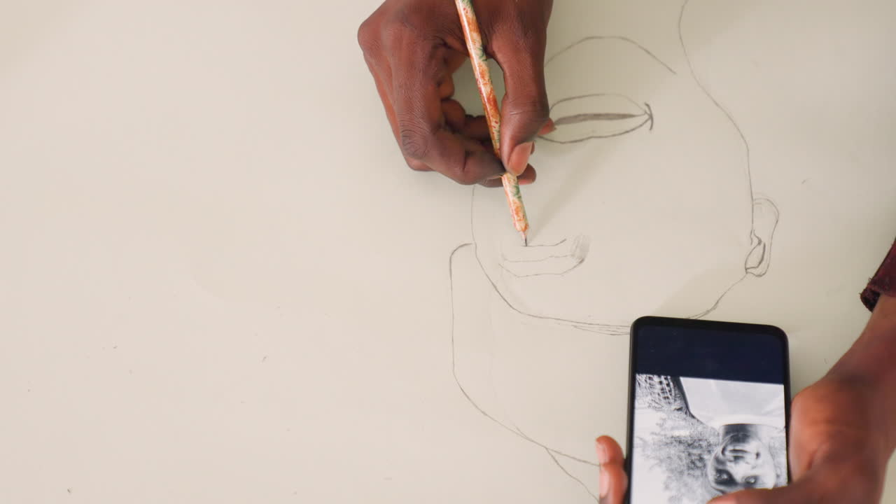 Artist finalizing portrait sketch with black painting reference on phone and pencil on paper. Wide composition shows hand refining eyes and mouth, subtle shading, intentional strokes and quiet studio