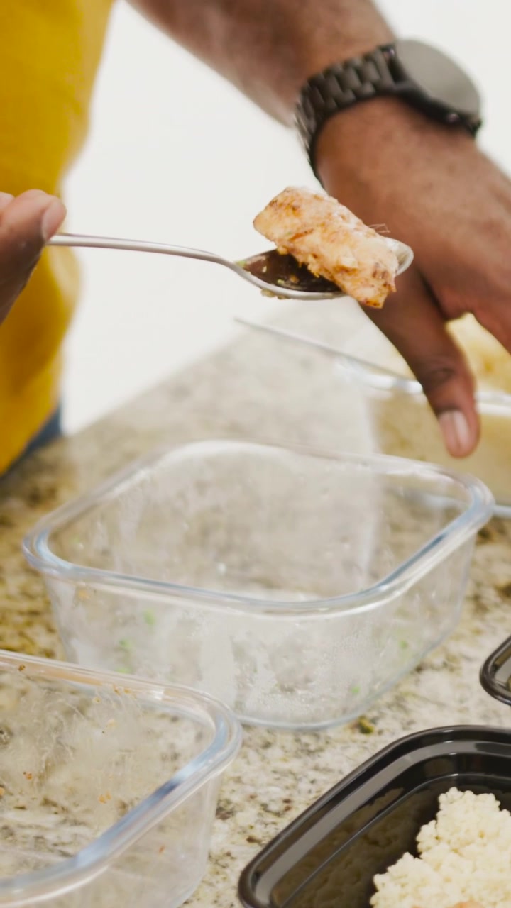 Cuoco che stabilizza un vassoio di riso nero, raccoglie il pollo con un cucchiaio, riempie contenitori di vetro per la preparazione dei pasti.