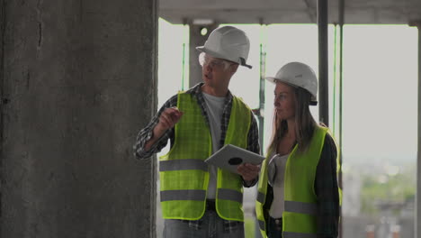 Zwei-Ingenieure,-Ein-Mann-Und-Eine-Frau-In-Weißen-Helmen-Mit-Einem-Tablet-Computer-Auf-Der-Baustelle-Verurteilen-Den-Bauplan-Des-Gebäudes