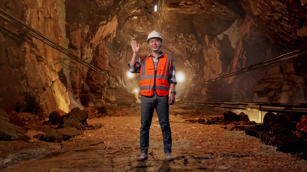 Full Body Of Asian Male Engineer With Safety Helmet Smiling To Camera And Waving Hand Saying Bye While Standing In Underground Mine Tunnel