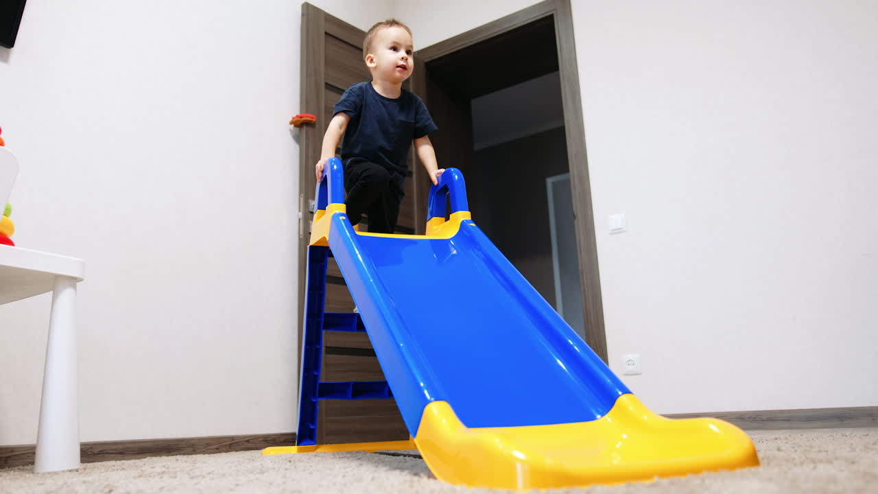 Caucasian toddler climbs on the slide and stops on the top. Kid slides down cheerfully smiling.