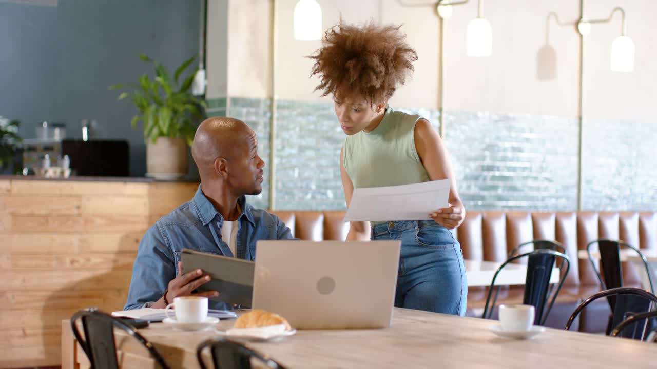 African American coworkers at cafe, woman showing paper, man pointing laptop and tablet, comparing