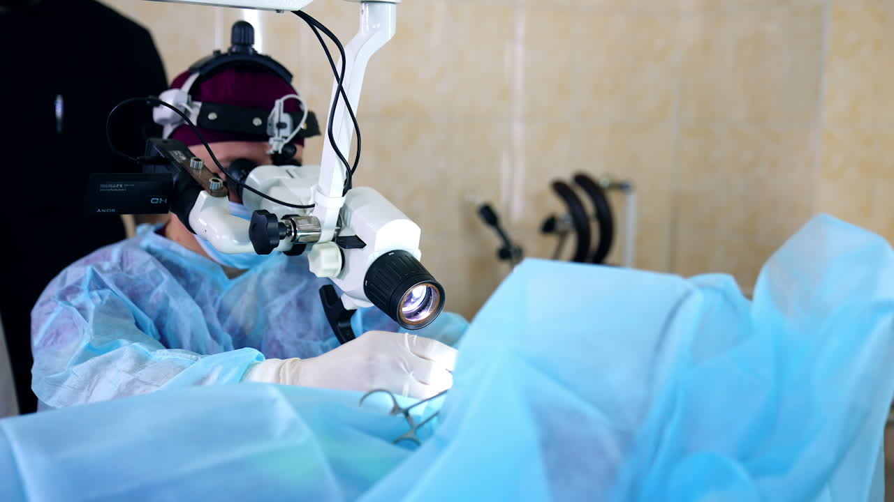 Doctor in blue uniform and mask sits looking at microscope performing medical procedure. Blurred backdrop.