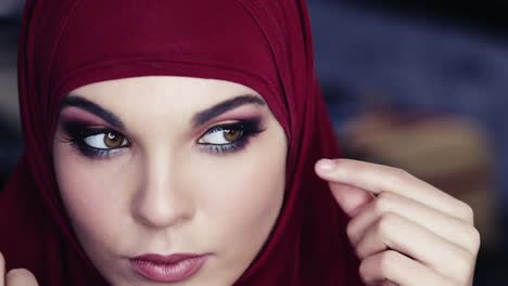 unbelievably beautiful girl with hazelnut eyes and purple hijab on her head is adjusting her traditional arabic head cover while cheking out her flawless make up